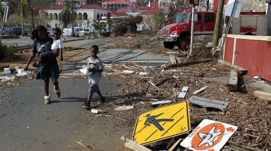 Žena s dvoma deťmi prechádza ruinami, ktoré...