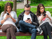 Group of friends two women and one man, sitting on a bench in park separately looking at their phones loosing communication. people using their phones and sending texts as they stand beside each other